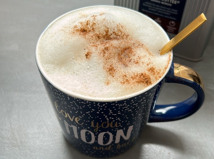 blaue Henkeltasse mit Milchschaum gefüllt. Ein goldener Löffelstiel schaut aus dem Milchschaum heraus. Der Schaum ist mit braunem Pulver dekoriert.