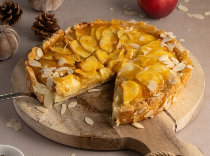 Runder Apfelkuchen mit Mandelblättern auf einem Holzbrett.