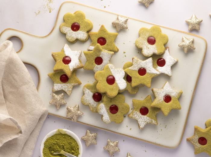 Hellgrüne Matcha-Mürbeteigplätzchen mit einem roten Marmeladenkreis in der Mitte und Puderzuckerdeko auf einem weißen Brettchen.