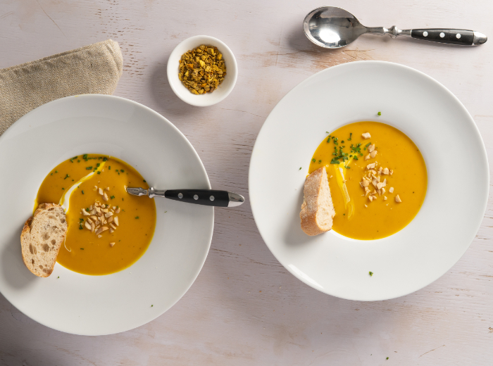 zwei runde weiße Teller mit orangefarbener cremiger Suppe und Baguettescheiben. daneben steht ein weißes schälchen mit losem Ingwer-Kurkuma-Tee.